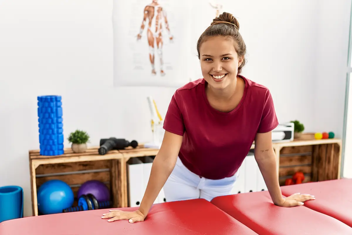 Junge Physiotherapeutin lächelt freundlich in einer modernen Praxis mit blauer Therapieliege und Sportgeräten im Hintergrund.