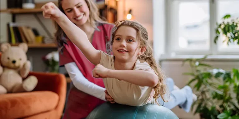 Physiotherapeutin unterstützt ein fröhliches Kind bei Übungen auf einem Gymnastikball in einer kindgerechten Umgebung.