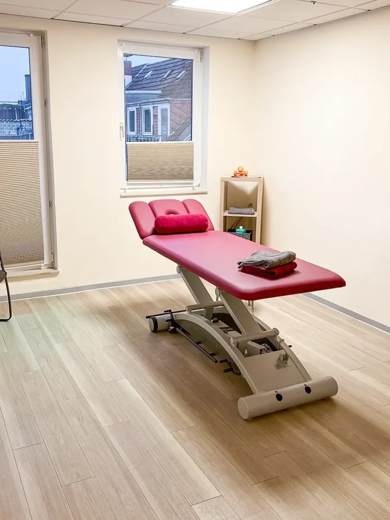 Behandlungsraum der Physiotherapie Mobili in Bremen mit roter Therapieliege, hellem Holzfußboden und großem Fenster.