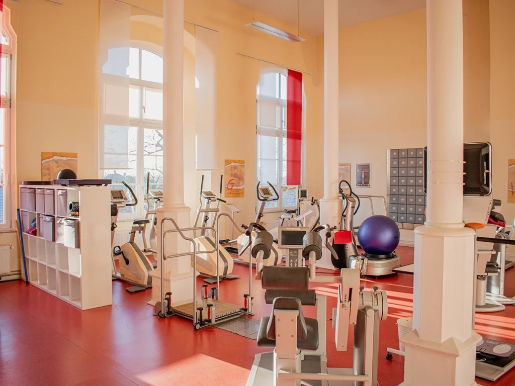 Großer Trainingsraum der Physiotherapie Mobili Celle mit roten Böden, Fitnessgeräten, großen Fenstern und weißen Säulen.