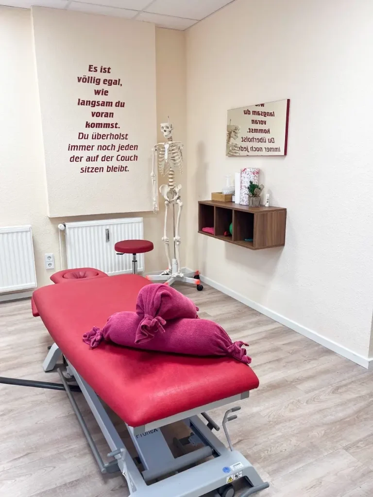Behandlungsraum der Physiotherapie-Praxis Celle mit roter Therapieliege, Skelettmodell und motivierendem Zitat an der Wand.