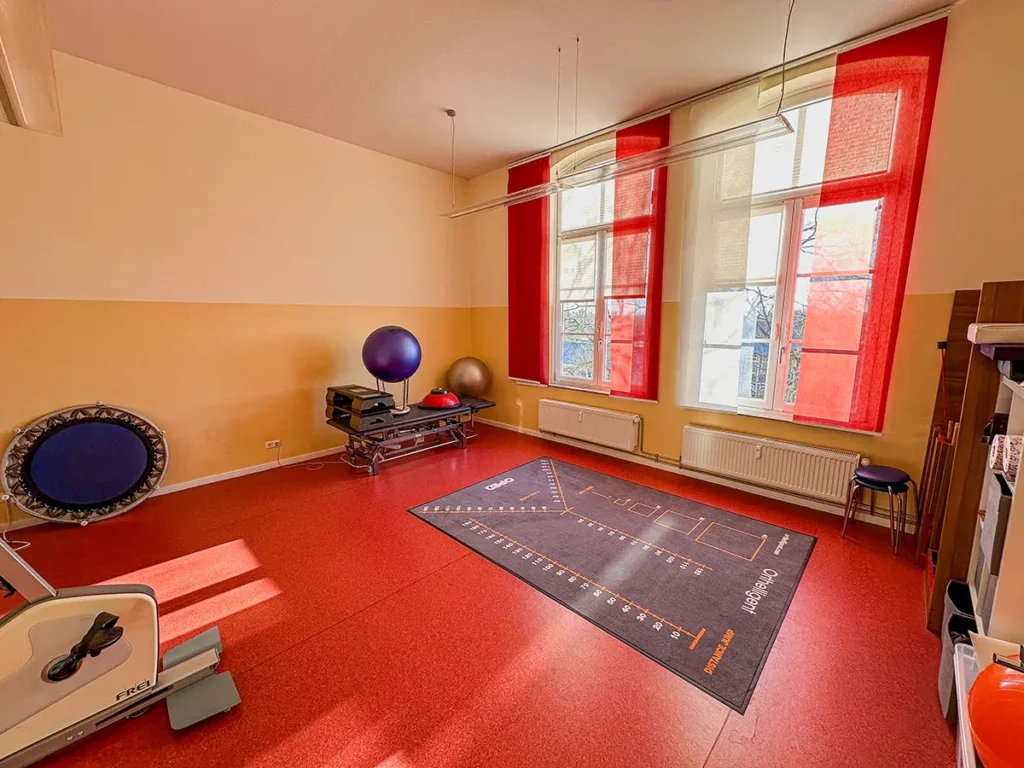 Trainingsraum der Physiotherapie Mobili Celle mit roten Bodenbelägen, großen Fenstern mit roten Vorhängen, Trampolin und Trainingsmatte.