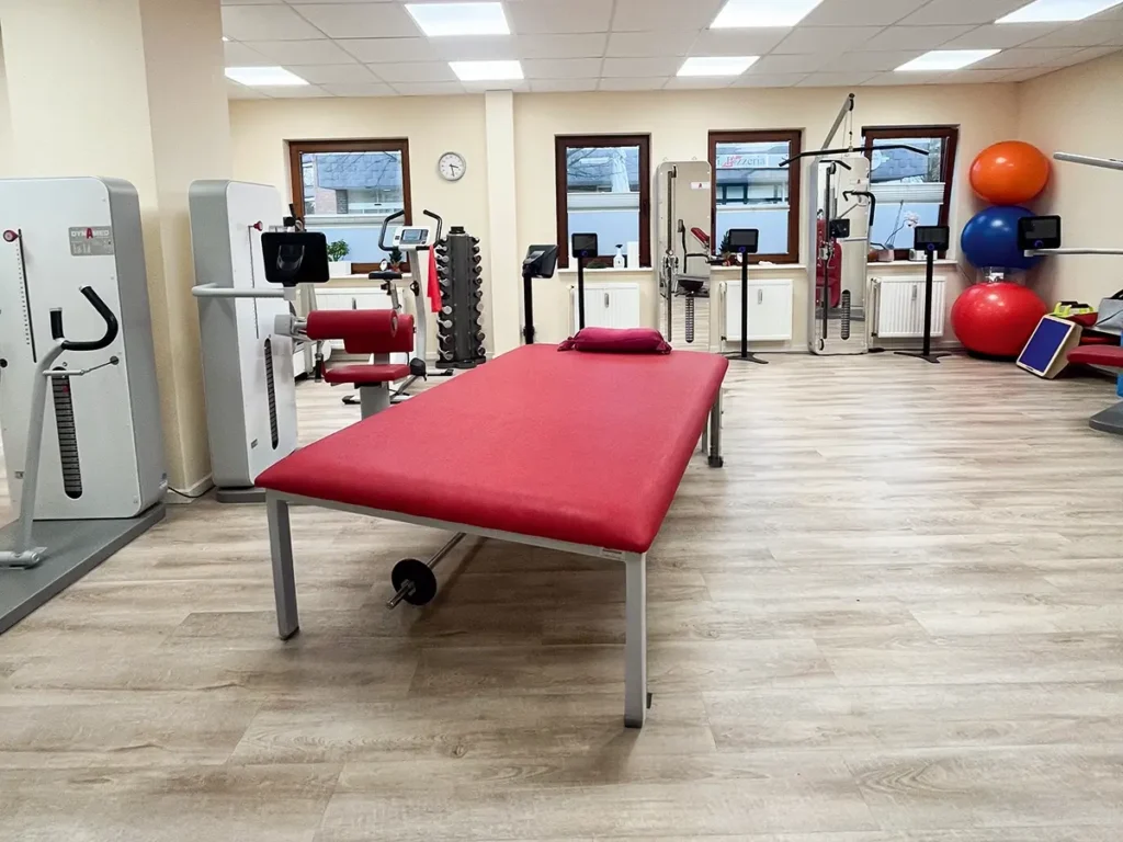 Moderner Trainingsraum der Physiotherapie-Praxis Celle mit Fitnessgeräten, roter Therapieliege und Gymnastikbällen.
