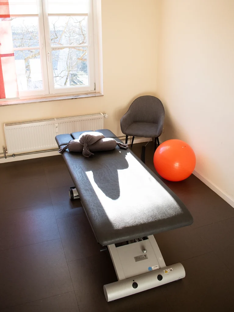 Behandlungsraum der Physiotherapie Mobili Celle mit einer Behandlungsliege, einem grauen Stuhl und einem roten Gymnastikball.