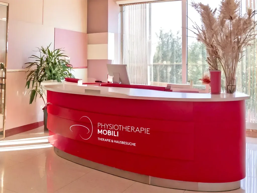 Empfangsbereich der Physiotherapie Mobili in Linden mit moderner roter Rezeption, dekorativen Pflanzen und natürlichem Lichteinfall.