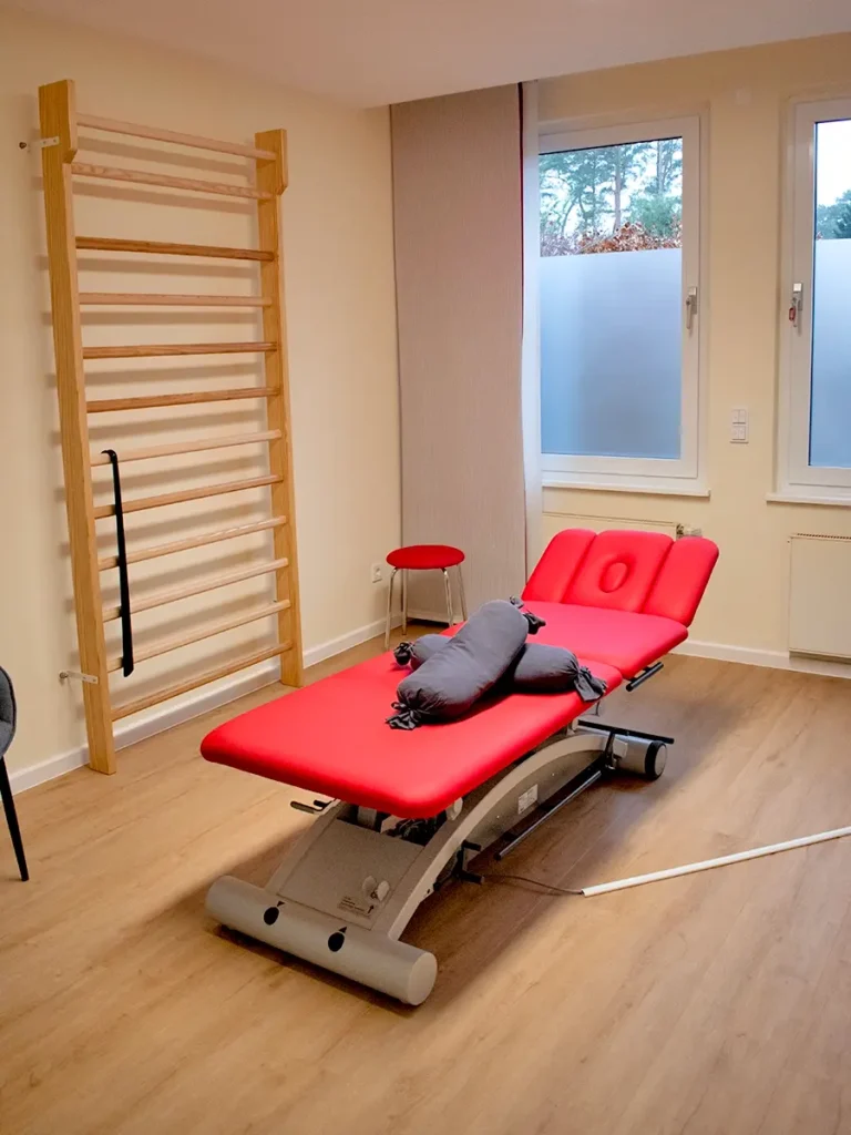 Behandlungsraum der Physiotherapie-Praxis Linden mit roter Therapieliege, Sprossenwand und modernem Holzfußboden.