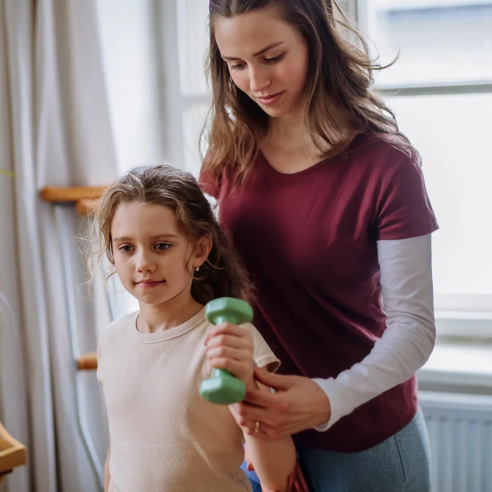 Therapeutin unterstützt junges Mädchen bei einer physiotherapeutischen Übung mit einer kleinen Hantel.