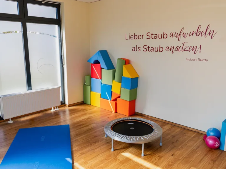 Heller Therapieraum mit bunten Bausteinen, Trampolin und Yogamatte für Kinderphysiotherapie.